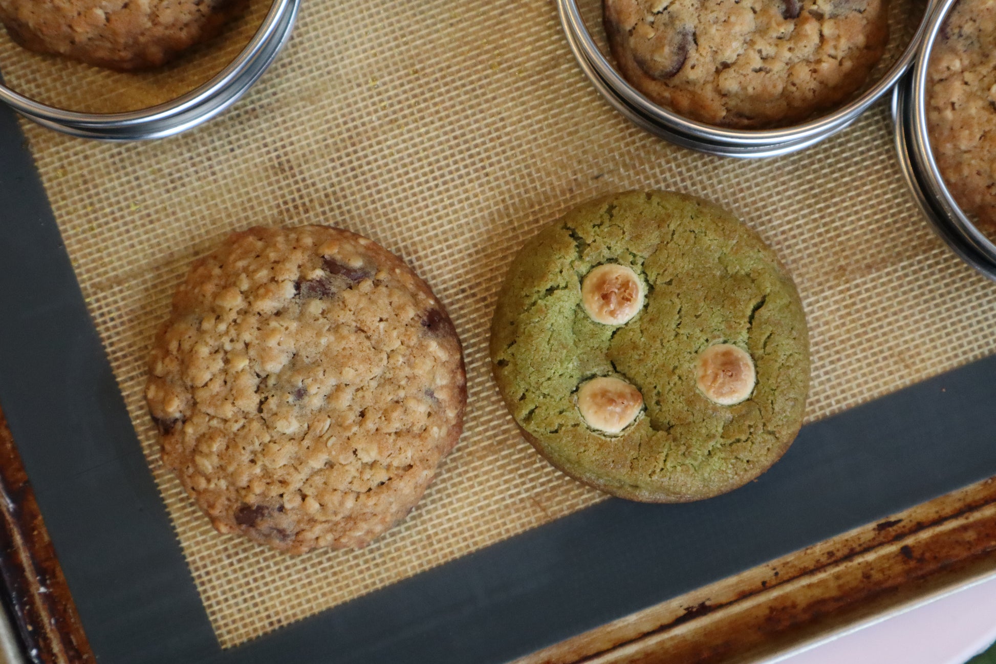 Two freshly baked cookies on a silicone baking mat: a chocolate chip cookie on the left and a green cookie with three mini marshmallows on the right, with metal baking cups containing more cookies at the top.