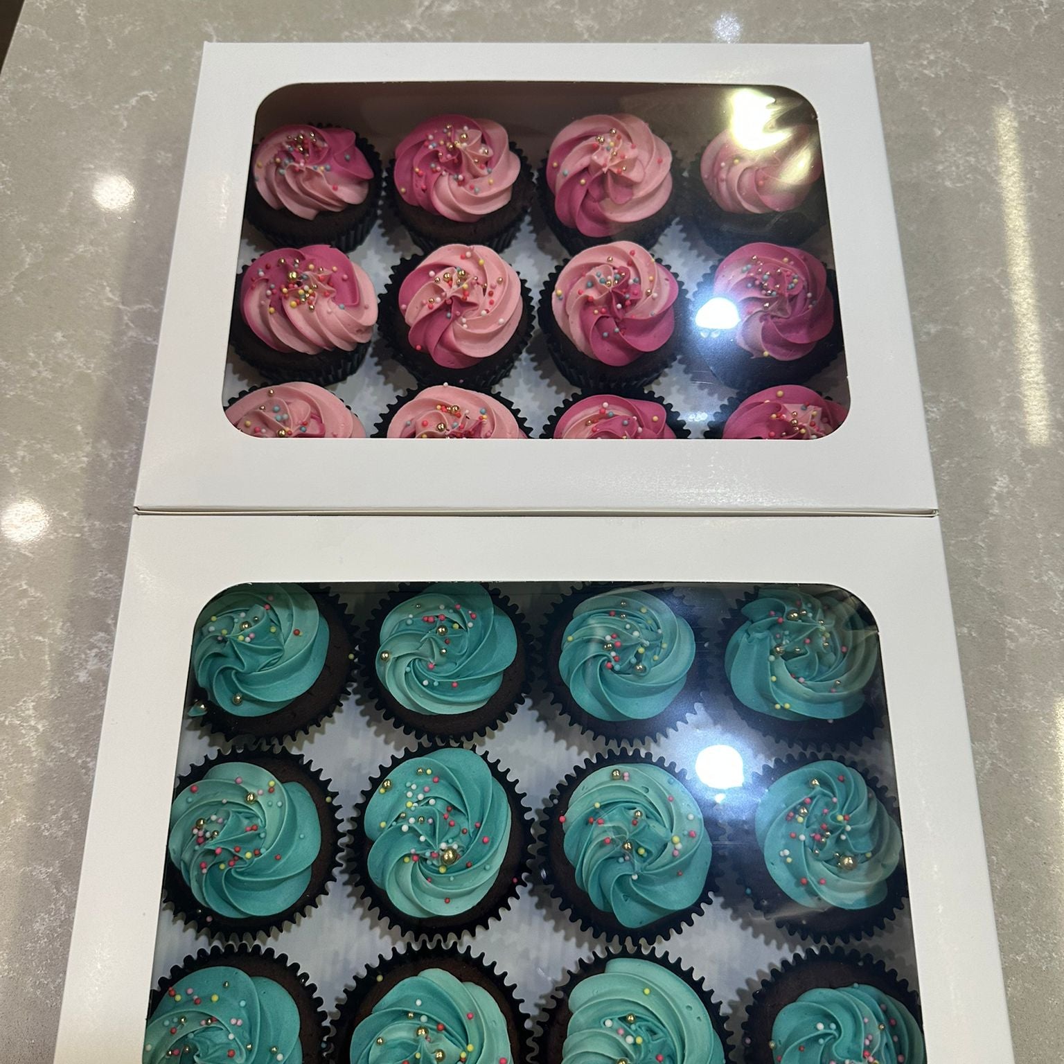 Two boxes of cupcakes with pink and teal frosting on a reflective surface.