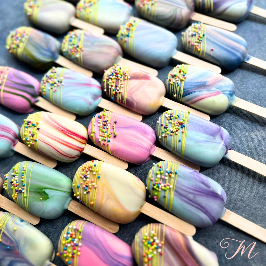 A selection of colorful eggless cakesicles with marbled designs and sprinkles on sticks.