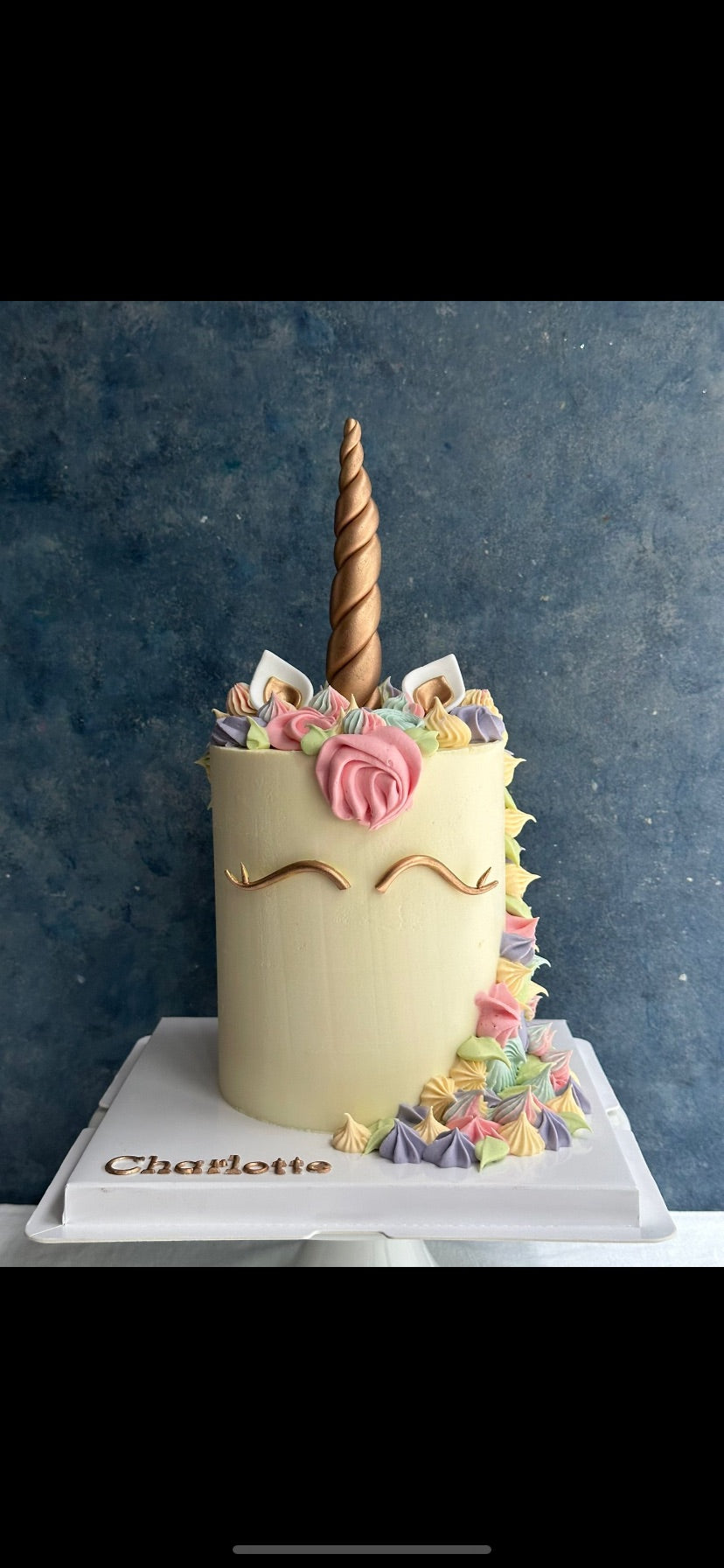 A custom unicorn-themed birthday cake with pale cream frosting, a gold horn, and pastel multicolor piped decorations, displayed on a white cake board with the name 'Charlotte' written on it.